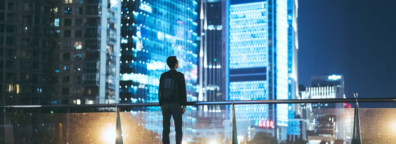man standing on balcony looking at lighted buildings of cityscape