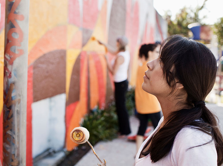 A person holding a paint roller and looking at a mural with other painters in the background