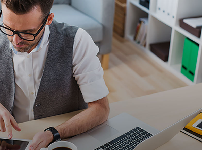 Person in a waistcoat looking down at a tablet with coffee and a laptop in the foreground
