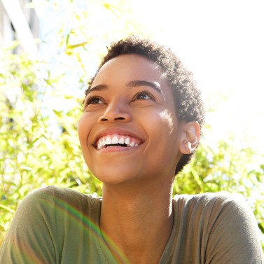 A woman sits in front of an area of greenery on a sunny day. She is relaxed and smiling. 