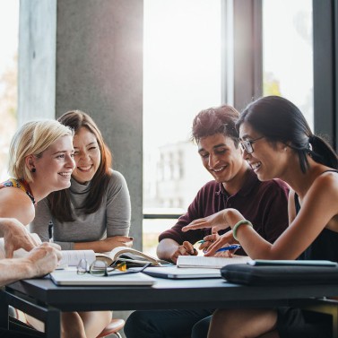 A group of young people of various ethnicities and genders sit around a table studying together. They are relaxed and smiling. 