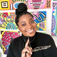 Vanessa Bellau, smiling towards the camera, holding a pair of glasses, in front of a bright colourful backdrop