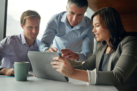 Image of three light skinned professionals, one woman, two men in an office looking at the screen of a mobile tablet