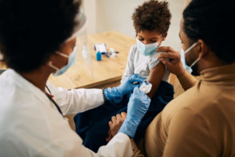 A brown skinned female doctor with afro hair wearing a mask and blue rubber gloves holding a needle about to inject a young toddler in the arm sitting on a man's lap, both brown skinned and wearing masks