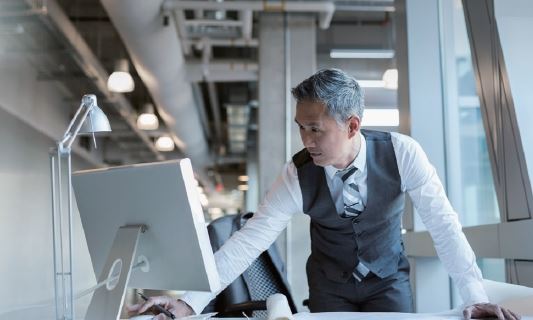 Image of a professionally dressed business man standing at a desk working from a computer screen in an industrial looking office 