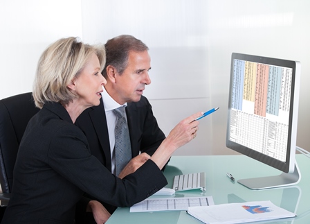 Image of a light-skinned middle aged woman and man in business attire sitting at a glass desk with papers on, look at a spreadsheet on a computer screen. The woman is pointing at the screen with a pen