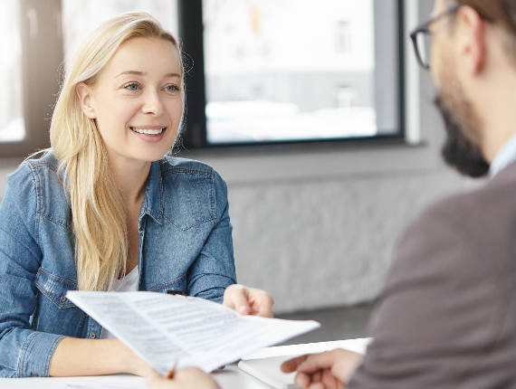 A casually dressed light skinned smiling blonde-haired woman sitting at a desk in front of a window, hands a printed document to a light skinned man in a suit, in profile with a beard and glasses sitting across the table