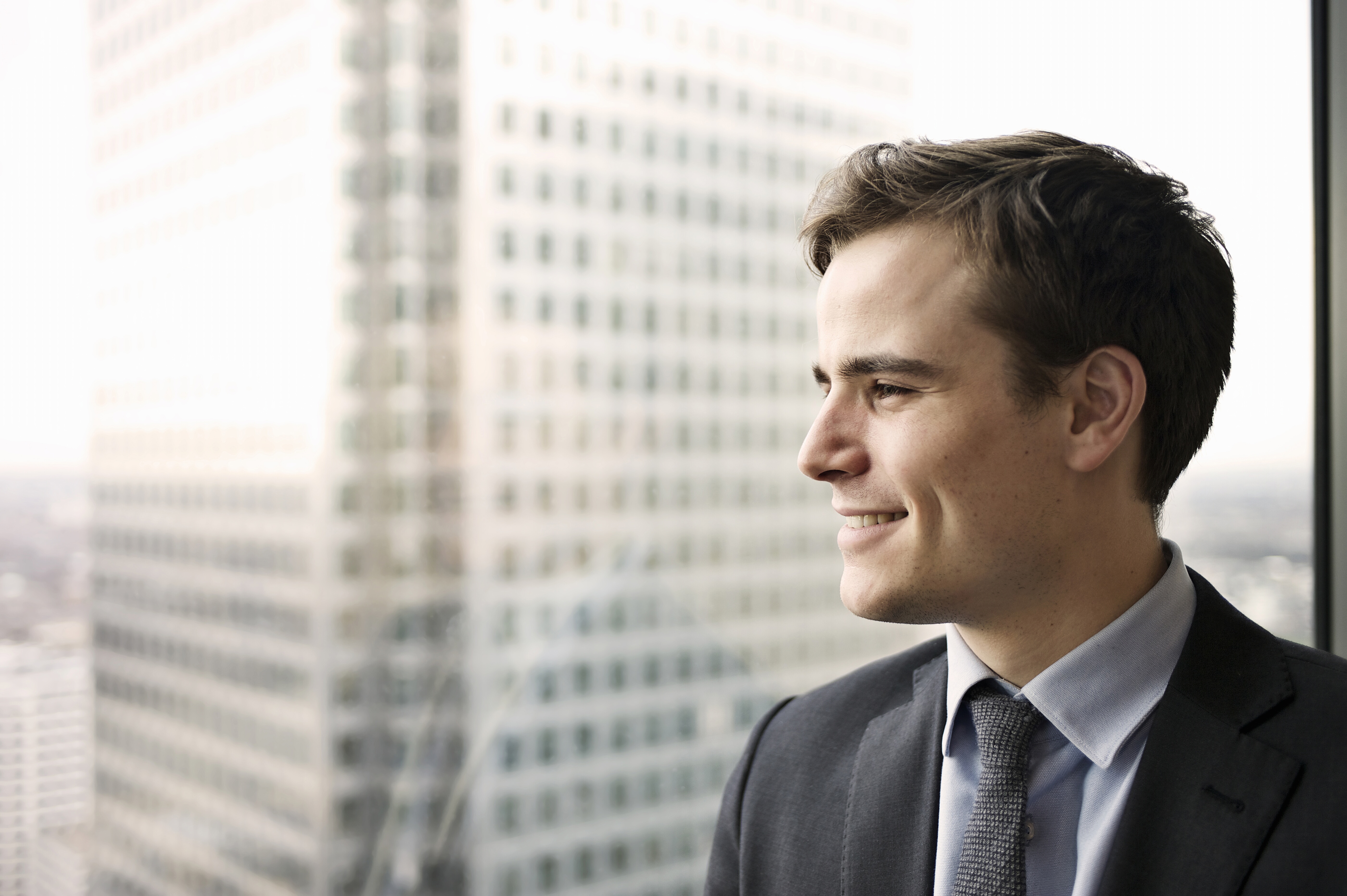 Portrait of a young business man smiling and looking out of a large window he is standing next to