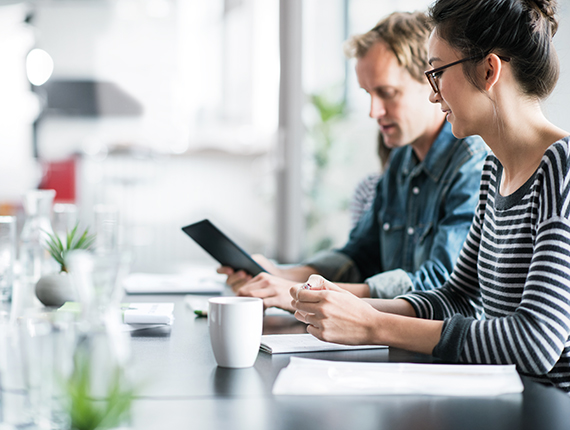 A casually dressed man with short blond hair and woman with dark hair pulled into a bun sitting at a table in a bright naturally lit meeting room. The man is holding what looks like a tablet which they are both looking at