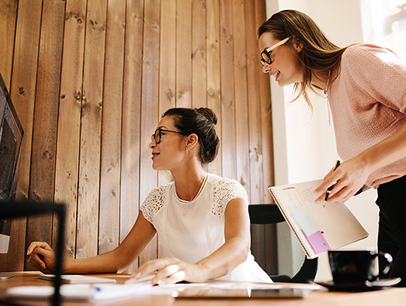 Two light skinned women wearing glasses smiling while looking at a computer screen. One is sitting and the other standing holding a paper file and a pen