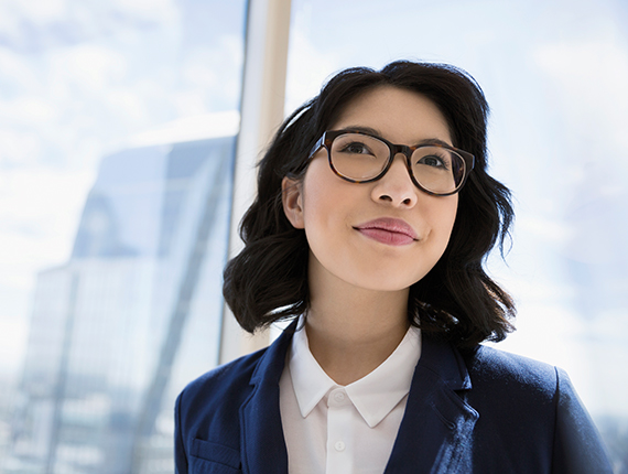 Image of a professionally dressed woman Asian woman wearing glasses, smiling and standing with her back to a large window in a bright sunny room