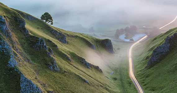 Scenic image of green hills and a valley with a road between showing a light trail from a car