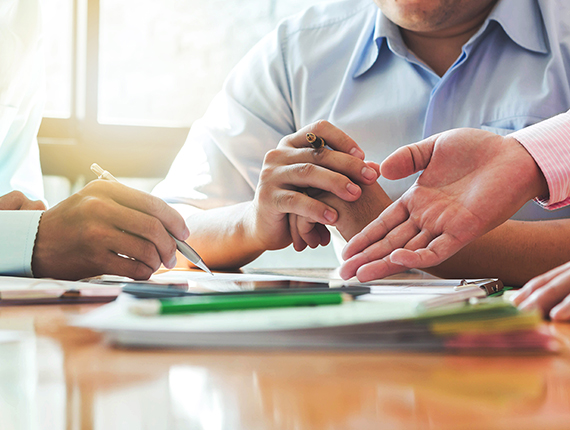 Hands of three men in focus. Man on the left is holding a pen in his left hand and pointing it at a piece of paper on the table, position in front of man in the middle. Man in the middle is wearing a blue shirt sitting at a table with his left hand resting on his right hand. Man on the right and pointing at the same piece of paper