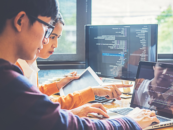 Two Asian professionals sitting in front of two computer screens working together on coding