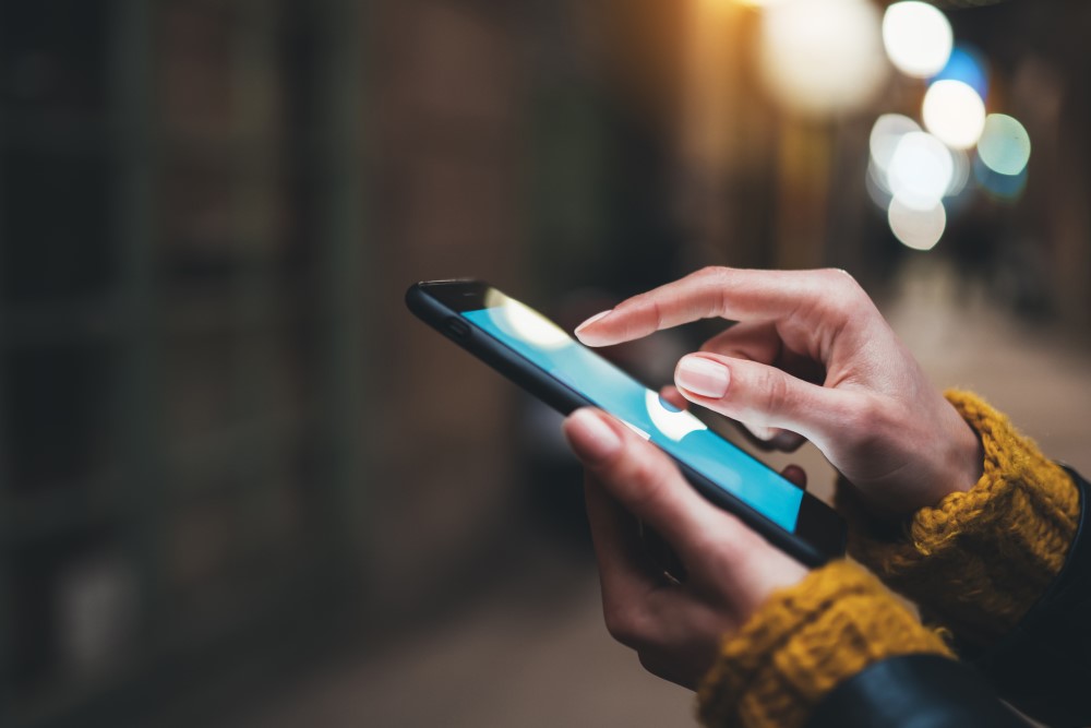 A blurred background. In focus are the sleeves of a mustard jumper and a pair of hands holding a mobile phone in the left palm while the right thumb and fore finger touch the phone screen