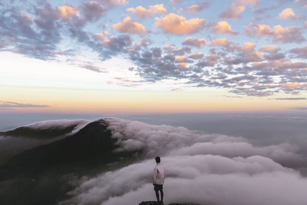 Man standing on a cloud covered mountain peak looking out at other cloud covered peaks 