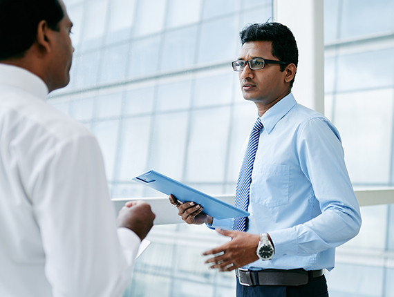 Head to waist shot of two Asian men standing in a bright room talking. The image shows the front of the man on right of picture in a blue shirt and striped blue tie holding a blue document holder with only a side view of the man on the left from his back in the frame