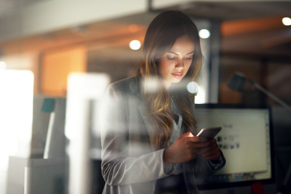 A young woman wearing a business suit standing by a window in an office looking at her mobile phone