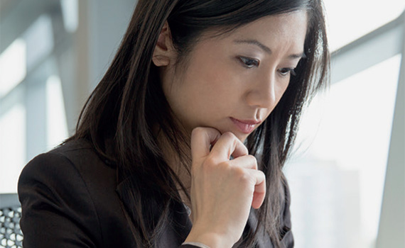 close up image of a head and shoulders of an Asian woman wearing a dark brown suit, sitting down in an office looking down at something in front of her