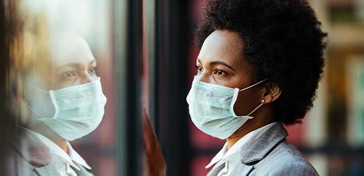 A black woman in a business suit with afro hair wearing a surgical face mask looking out a window where her face is reflected.