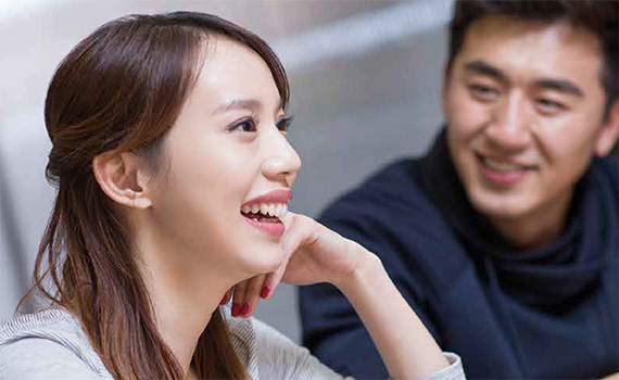 Head and shoulder shot of two casually dressed Asian looking man and woman smiling. The woman's head rest lightly against her left hand