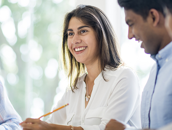 Head an shoulders image of two adults, a woman and man in business attire, in an office smiling. Woman is light skinned woman holding a pencil the man is brown skinned