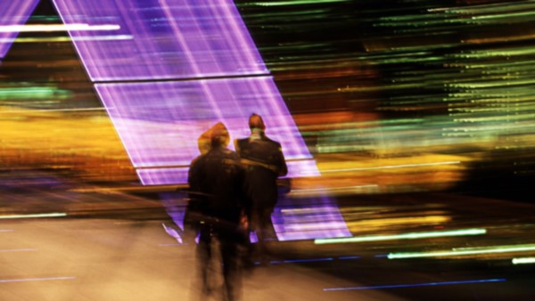 Image of a mixture of medium blurred colours in horizontal lines, and two people also blurred in front of three left to right slanted violet stacked blocks