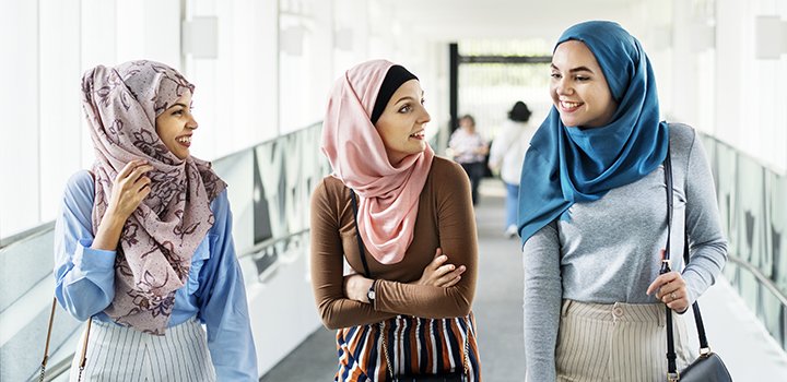 Image of three light-skinned young women with head coverings (likely hijab), smiling in mid-conversation standing in a bright hallway s