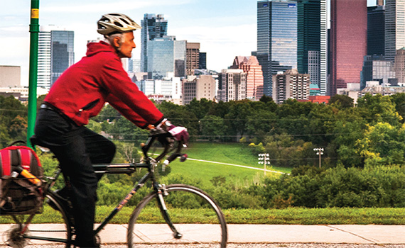 Image of a older looking man on a bicycle in the foreground dressed in a red outer jacket and helmet. The background is a city skyline of skyscrapers and trees 