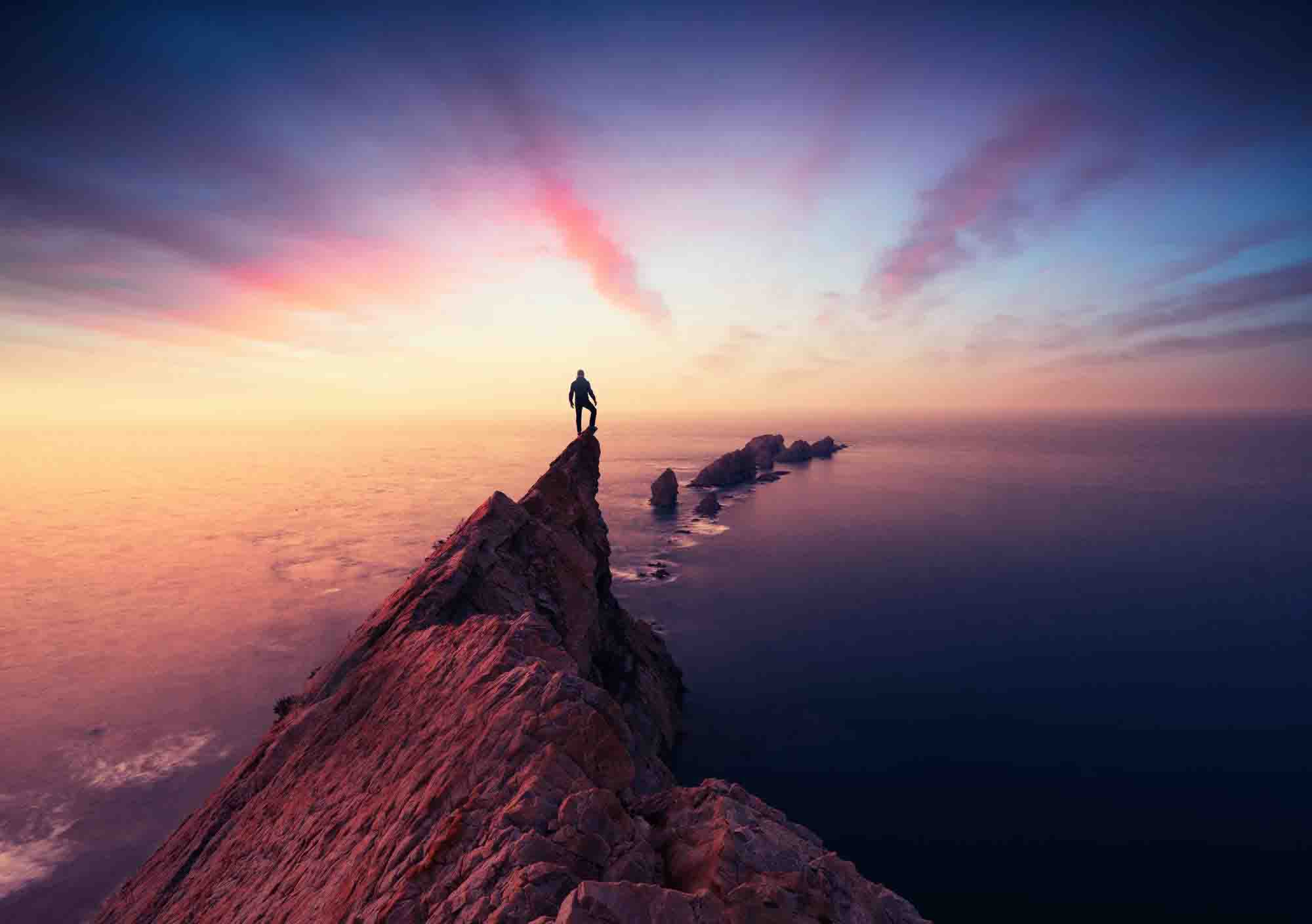 Image of a person standing on top of a mountain peak at sunset looking out at sea