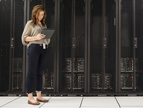 image of a casually dressed light skinned woman with wavy hair standing in front a wall of computer servers, looking at a mobile tablet screen she is holding