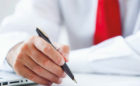 Image of mid section of a man in the background wearing a white shirt and red tie. In the foreground his right hand is writing with a pen