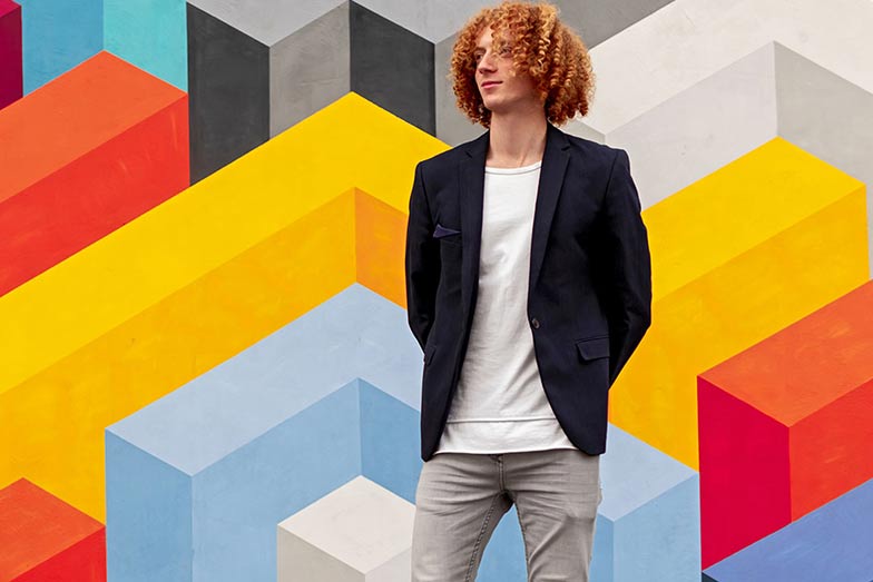 man with long curly hair standing in front of colourful blocks