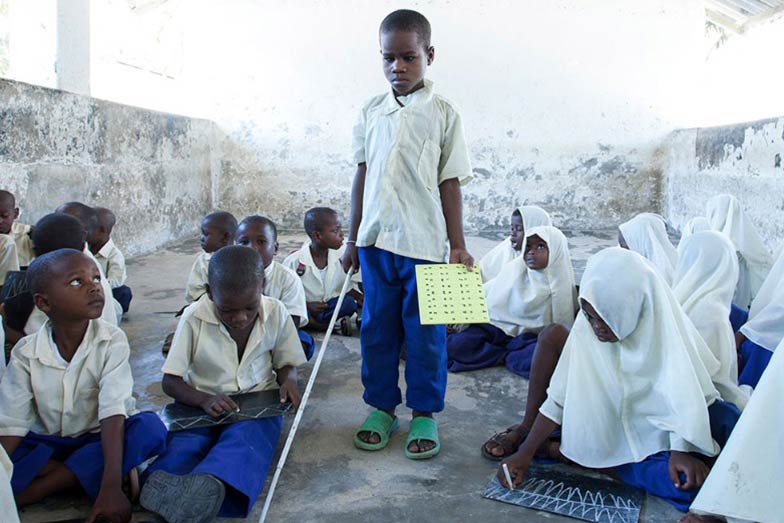 child with cane standing in a classroom with students sitting on the floor