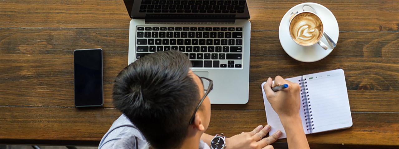 aerial view of a person writing on a notepad next to a laptop and a coffee