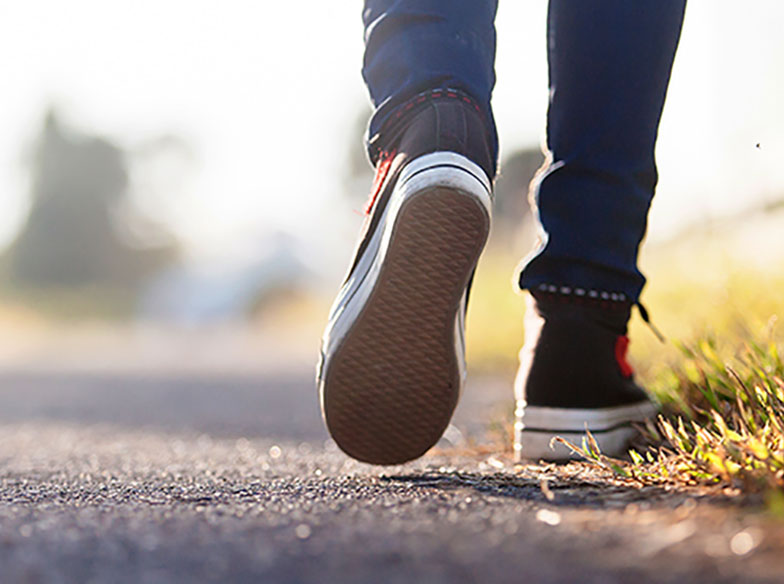 Back view of a person's feet in trainers walking on a sunny road