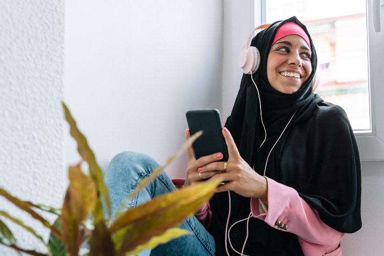 happy young person wearing hijab and headphones listening to media from mobile phone