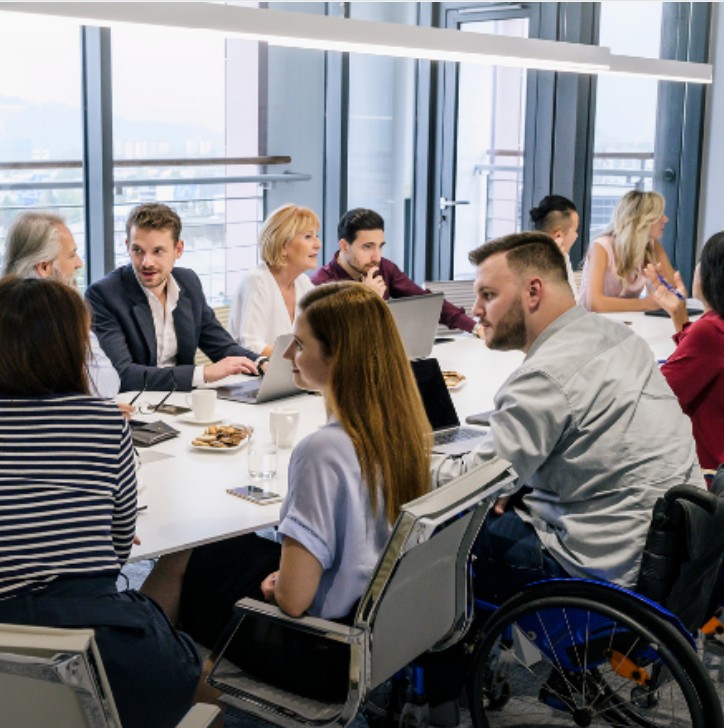 An image of a group of colleagues, including a wheelchair user, seated around a long table in a meeting. 