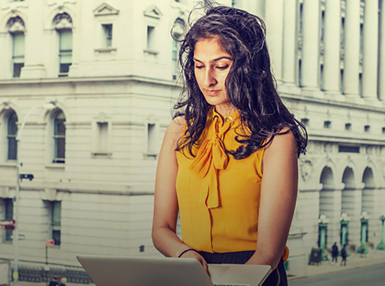 Person in a yellow shirt looking down at a laptop in front of a white building