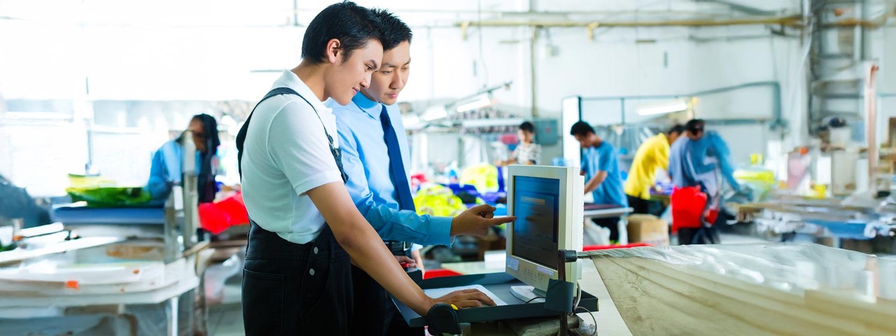 two persons looking at a computer screen with a factory in the background