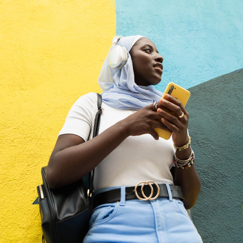 Young african person wearing hijab and earphones holding mobile phone in front of colourful wall
