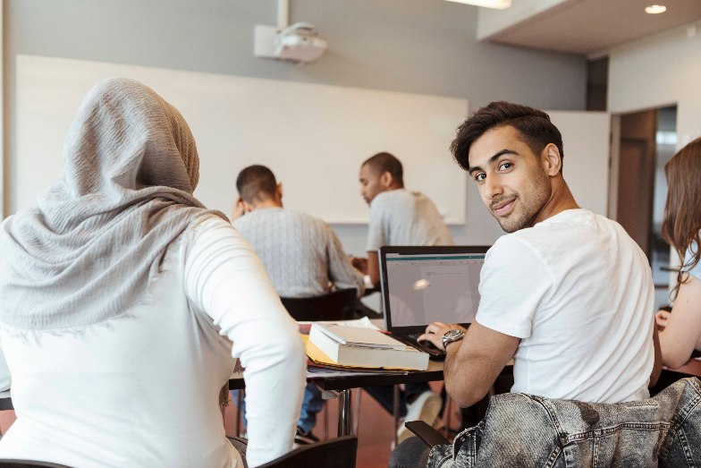Student in classroom on laptop looking over his shoulder to smile at camera