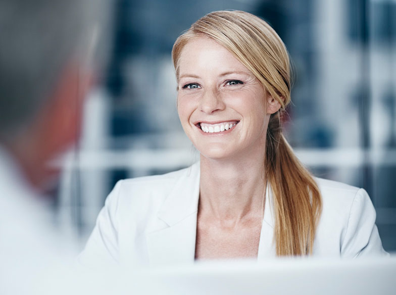 Person in an office smiling face-on to camera with rear view of a person's head in foreground
