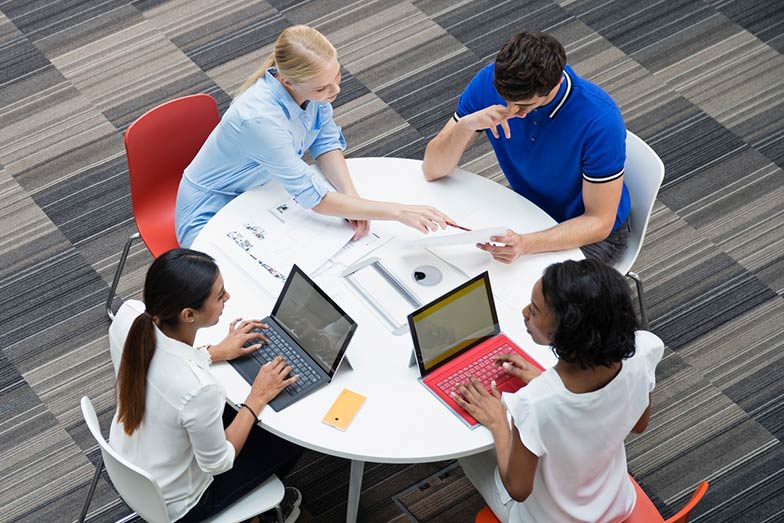 four persons sitting at a table with laptops and books
